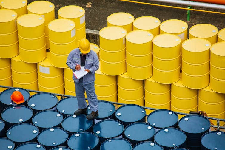 A worker stands atop oil barrels with a clipboard.