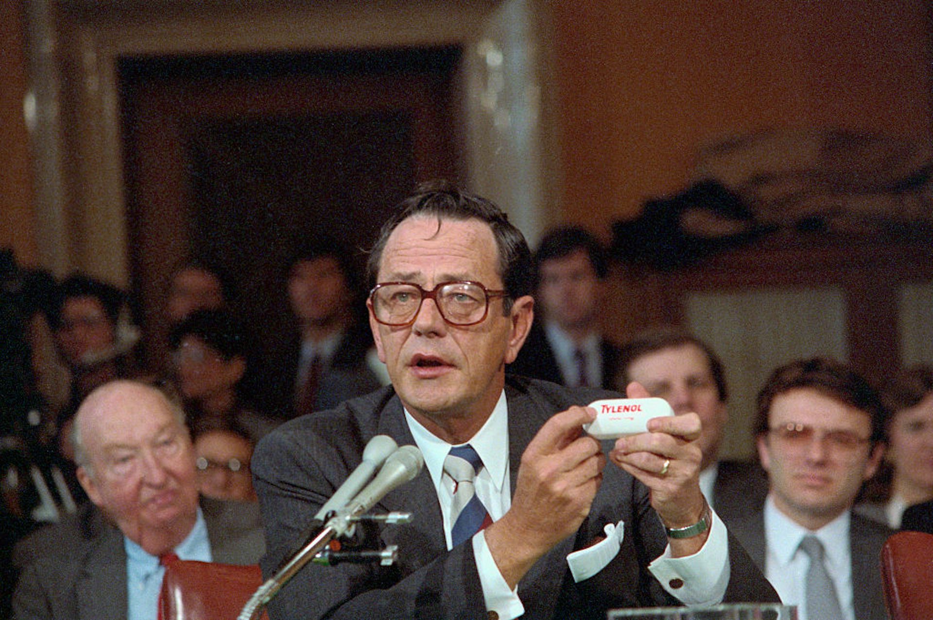 Man wearing glasses and suit speaks into microphone while holding a white bottle of pills.