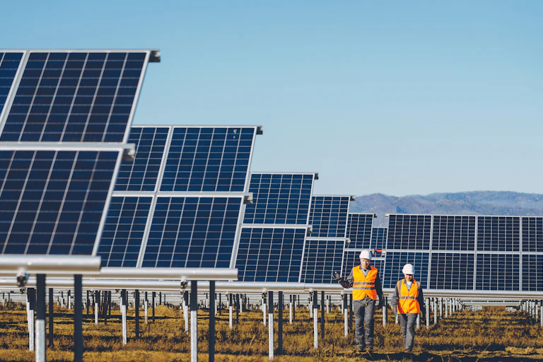 Two people in reflective orange jackets walking in front of a solar farm against a mountainous backdrop.
