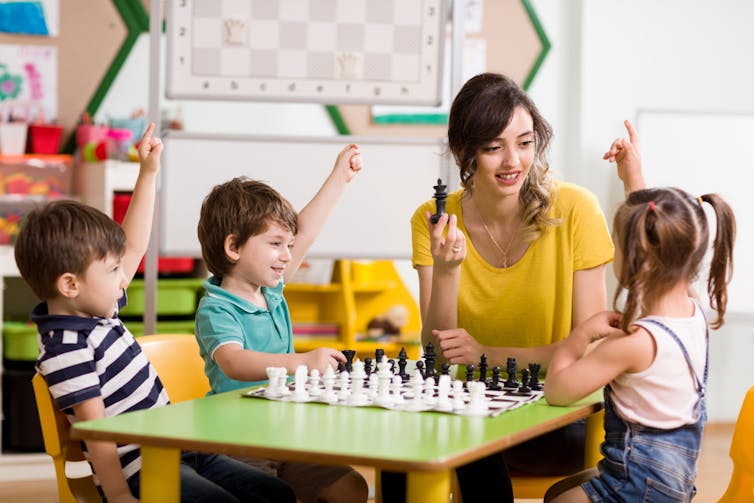 A woman and three young children sit around a chessboard table.  The woman holds a chess piece in her hand, while the children raise their hands.
