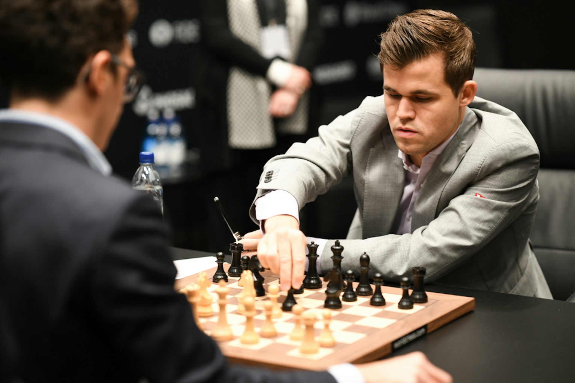 A man wearing a grey suit moves a piece on a chess board.