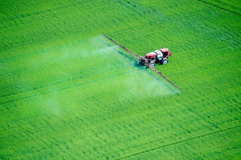 A vehicle with a large tank moves across a field, spraying a white substance behind it