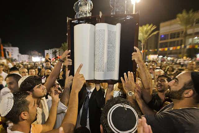 Men in a crowd gather around a large book held above their heads.