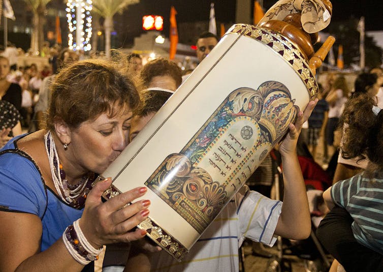 Una mujer con una camisa azul de manga corta besa un estuche adornado que sostiene un libro sagrado en medio de una multitud de personas.