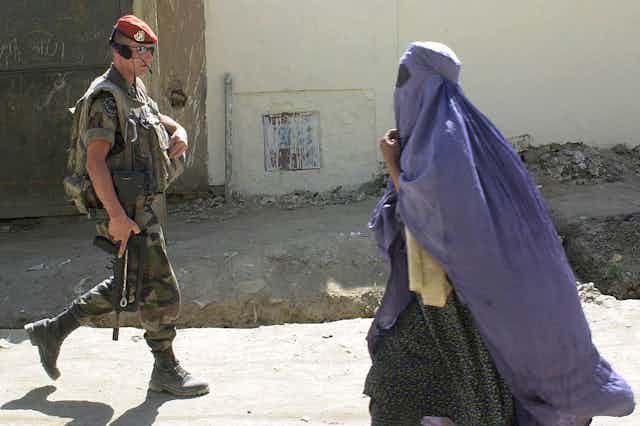 An Afghan woman in a purplish blue burqa walks past a French soldier wearing sunglasses and a maroon beret.