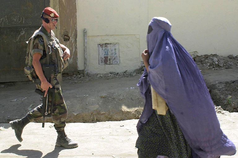 An Afghan woman in a purplish blue burqa walks past a French soldier wearing sunglasses and a maroon beret.