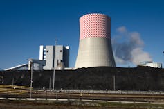 A coal-fired power plant with a large chimney in the centre in front of a large wall of coal deposits.