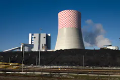 A coal-fired power plant with a large chimney in the centre in front of a large wall of coal deposits.
