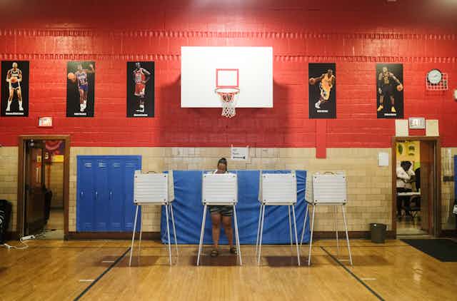 A woman standing behind a voting station in a gym.