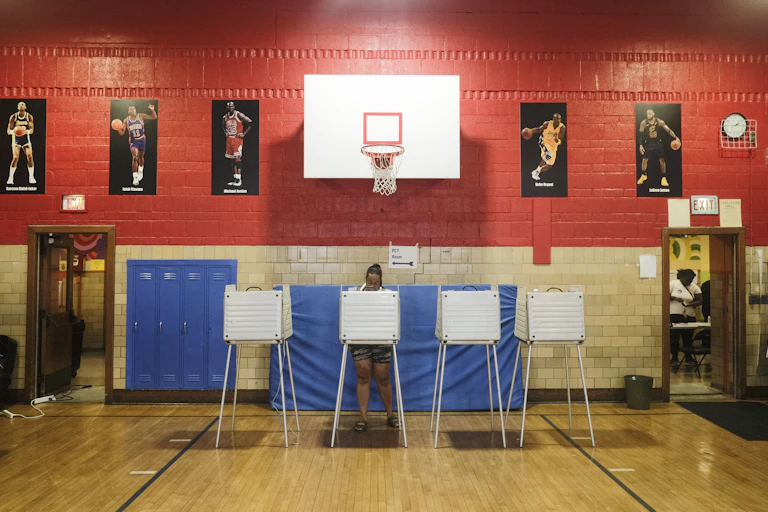 A woman standing behind a voting station in a gym.