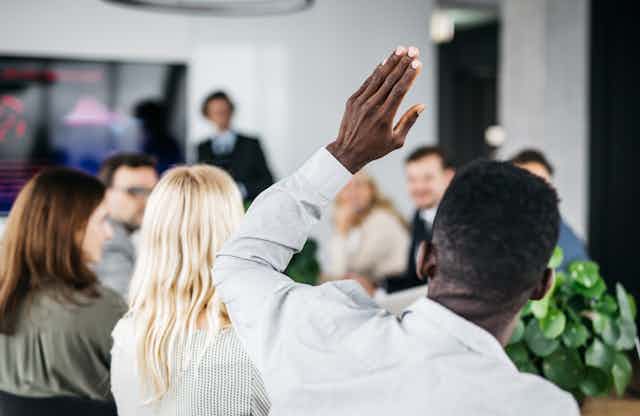 a black man raises his hand in a meeting with other people in the foreground