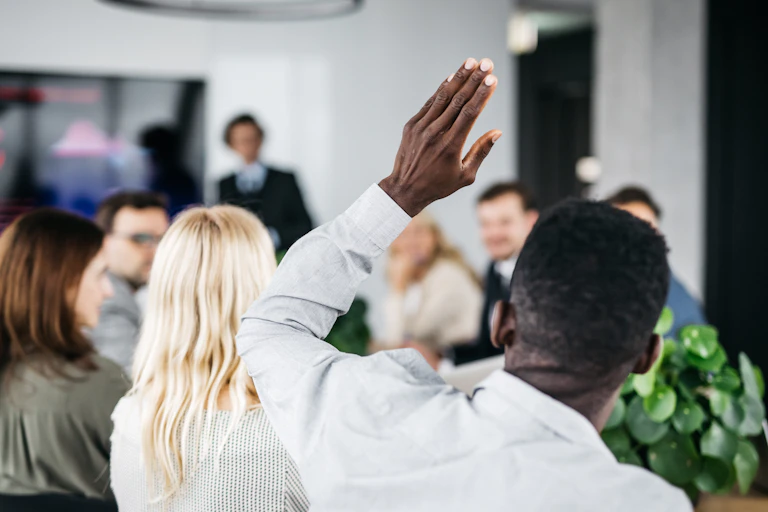 a black man raises his hand in a meeting with other people in the foreground