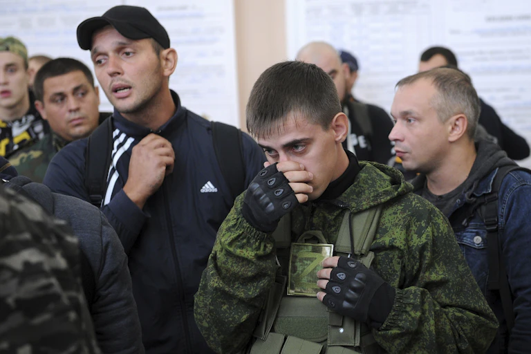 Young men stand together. The man in the foreground holds a card that displays the letter Z and has his hand over his nose and mouth.