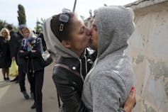 A woman in a leather jacket and a man in a grey hoodie kiss and embrace.