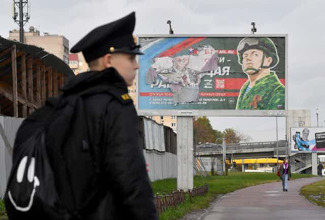 A young man in a black jacket and cadet like hat stands in front of a torn billboard with the drawing of a soldier in green camouflage and Russian words.