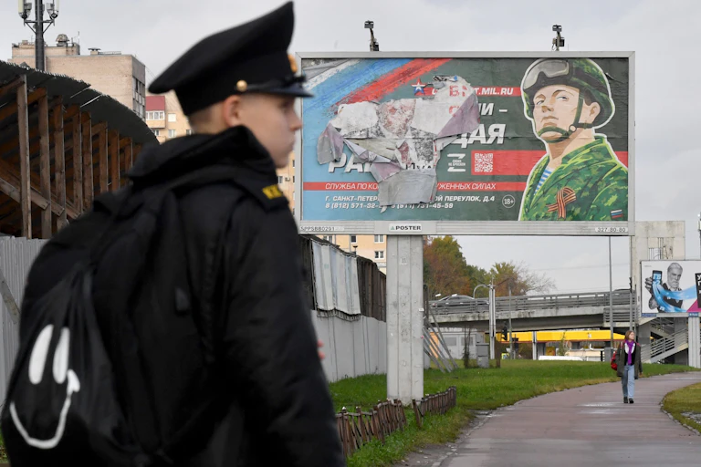A young man in a black jacket and cadet like hat stands in front of a torn billboard with the drawing of a soldier in green camouflage and Russian words.