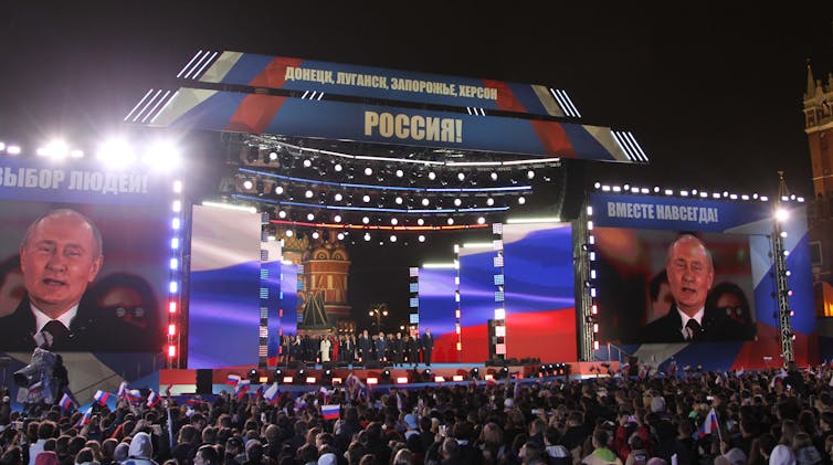 A large stage shows people from a distance, with two large screens on either side showing a middle-aged to older white man.