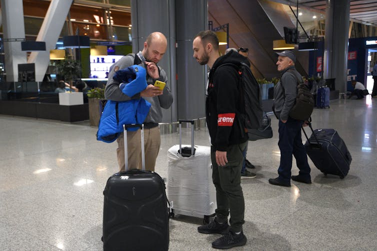 Two young adult men stand looking at a phone in an airport, with rolling suitcase at their sides.