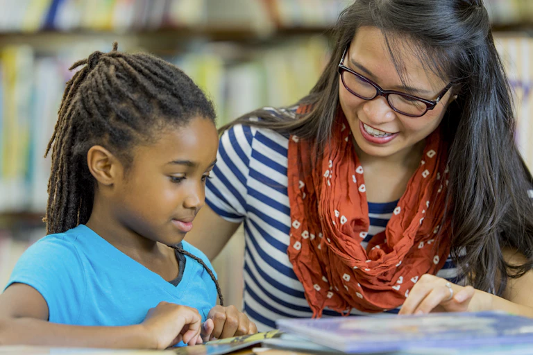 A young Black girl in a turquoise shirt reads a book while a woman in a blue and white striped shirt sits nearby.