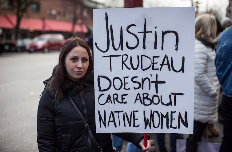 A dark-haired woman holds a sign that reads 'Justin Trudeau doesn't care about native women.'