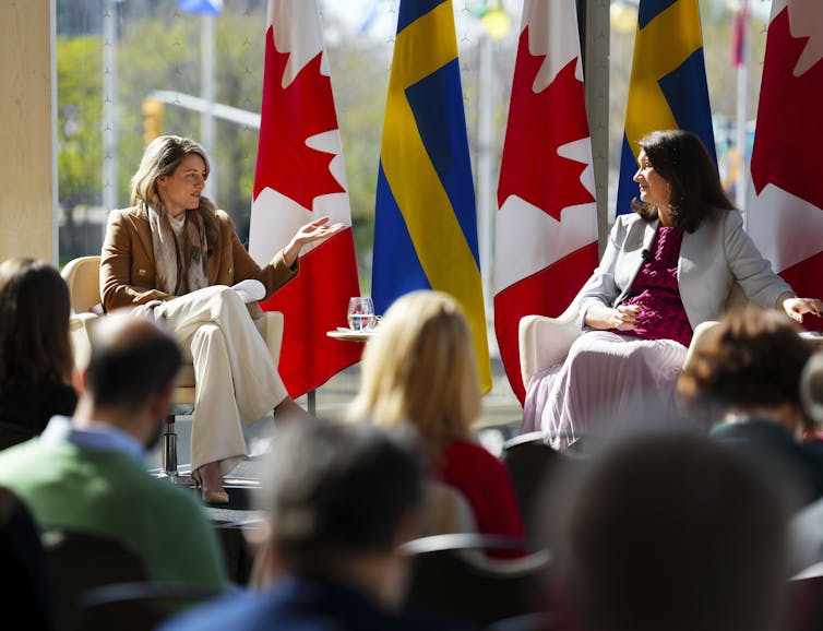a white blonde woman sits talking to a dark-haired white woman with the canadian and swedish flags behind them