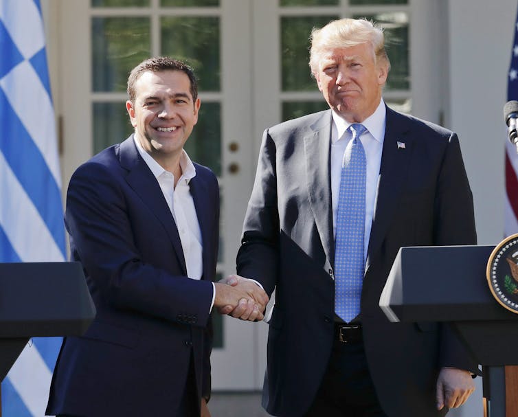 A blond aging man shakes hands with a younger dark-haired man.