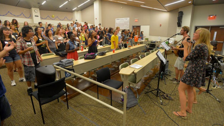 A crowd of students in a classroom, many of them with their hands up in worship, facing two singers at the front.