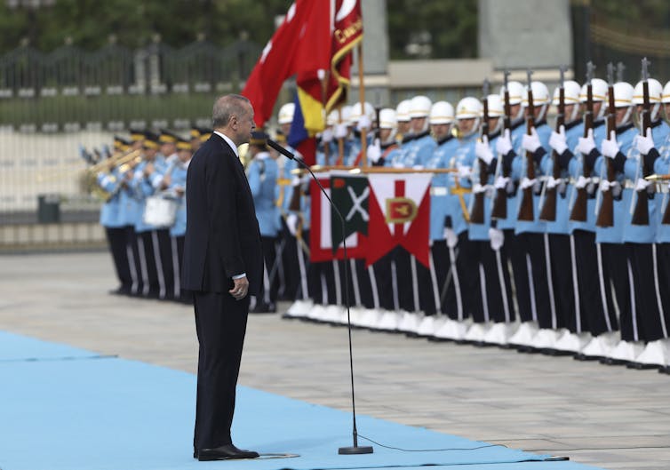 رجل يرتدي بدلة يقف على سجادة زرقاء شاحبة وينظر إلى حرس الشرف العسكري الذي يرتدي نفس الظل الأزرق.