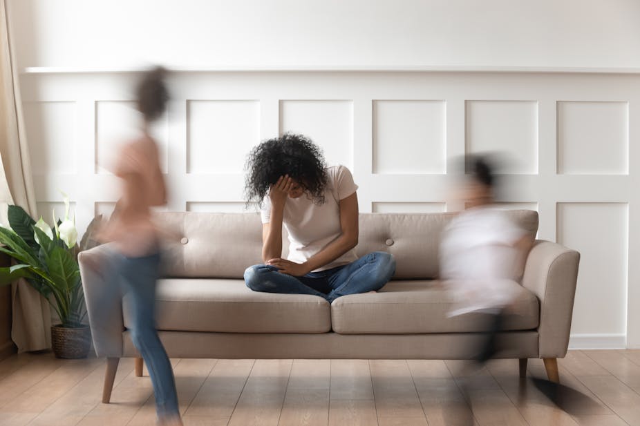 Young woman sits on a sofa, head in hands, while blurred images of children appear to move past her.