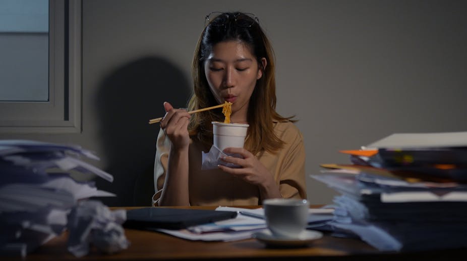 A young woman eats a pot noodle using chopsticks.