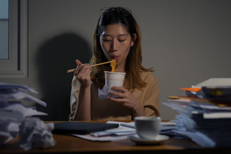 A young woman eats a pot noodle using chopsticks.