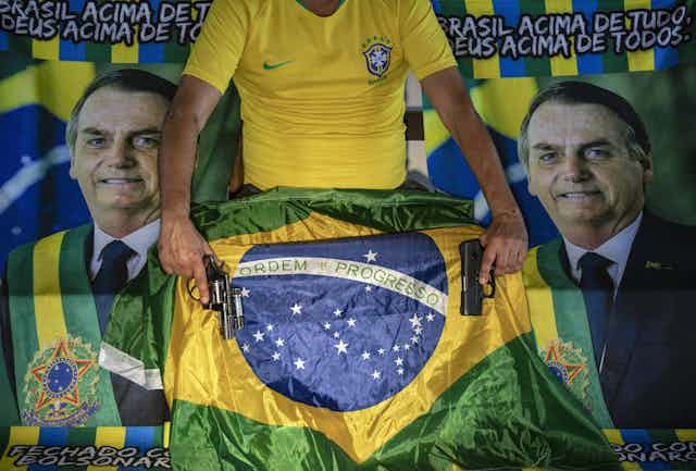 A man holds two guns in front of a Brazilian flag and pictures of Jair Bolsonaro.
