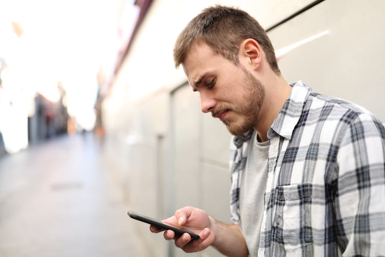 A young man in a plaid shirt looking at his phone with a worried expression.
