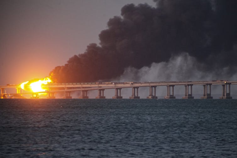 Flames and billows of black smoke trail above a bridge structure over a body of water.