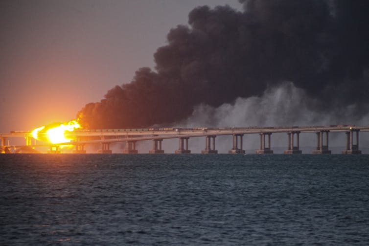 Flames and billows of black smoke trail above a bridge structure over a body of water.
