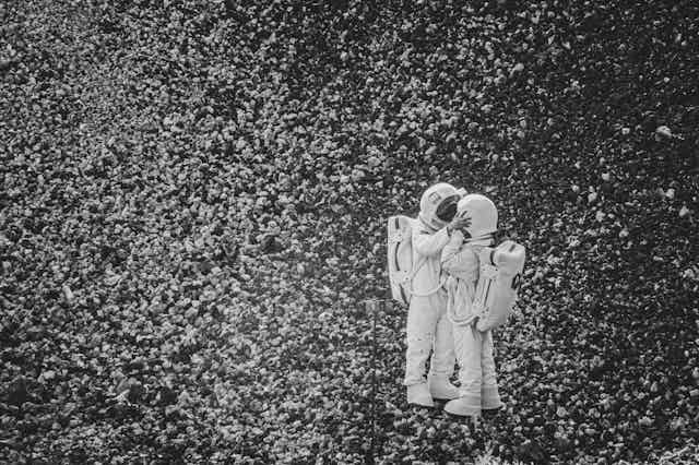 black and white photograph showing two people in astronaut costumes standing on a rocky landscape — one has their hands on the sides of the other's helmets