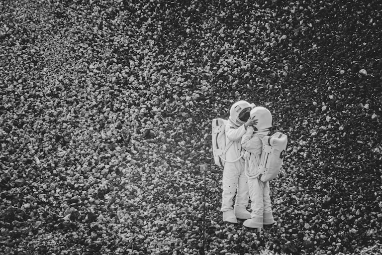 black and white photograph showing two people in astronaut costumes standing on a rocky landscape — one has their hands on the sides of the other's helmets