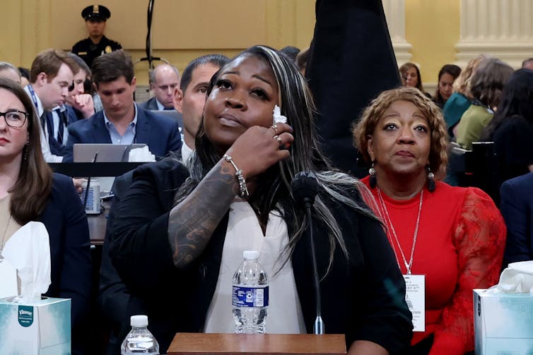 A woman dressed in a black jacket and white shirt wipes tears from her face while giving testimony at a table in a large room filled with people.
