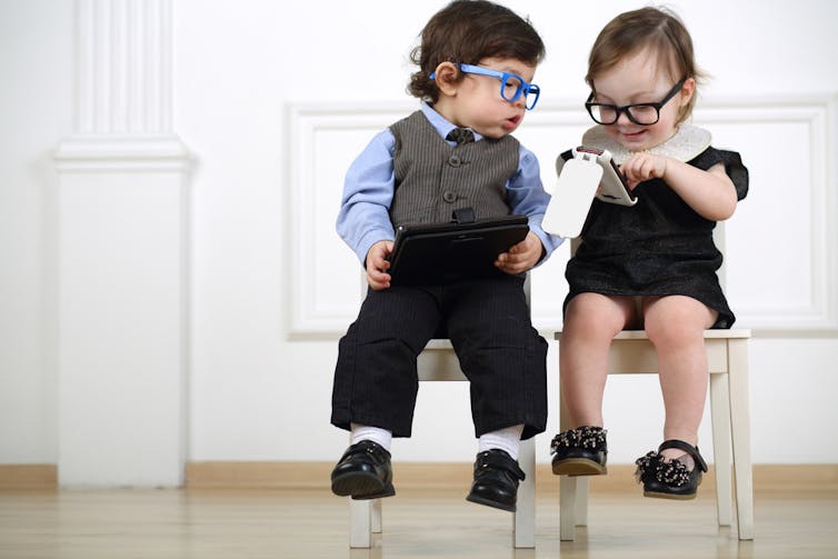 Two small children with glasses sitting on white chairs : a boy with a tablet computer, a girl with a cell phone