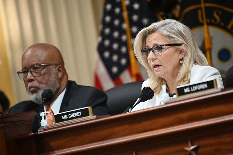 A man wearing glasses and in a dark suit, sitting in front of an American flag along with a woman in a white jacket and wearing glasses.