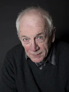 An elderly bald headed man in a dark shirt and jumper.