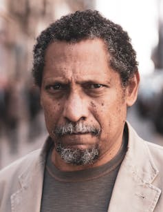 A slightly frowning, middle-aged black man in a tan jacket.