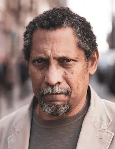 A slightly frowning middle-aged black man in a beige jacket.