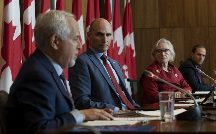 Three men in suits and one woman in a blazer sit behind a table. Behind them is a row of Canadian flags