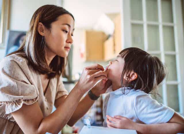 Young Asian mom inserts a nasal swab into her daughter’s nose while carrying out a COVID-19 rapid test at home.