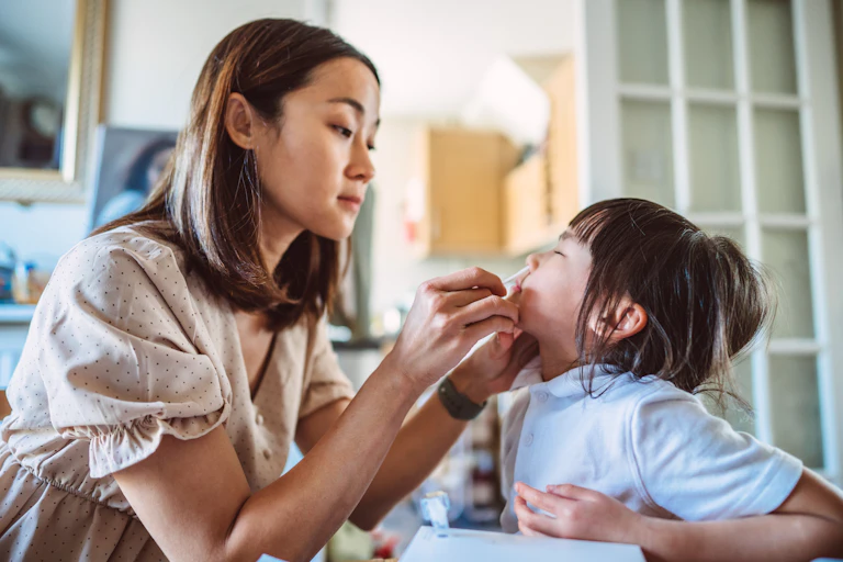 Young Asian mom inserts a nasal swab into her daughter’s nose while carrying out a COVID-19 rapid test at home.