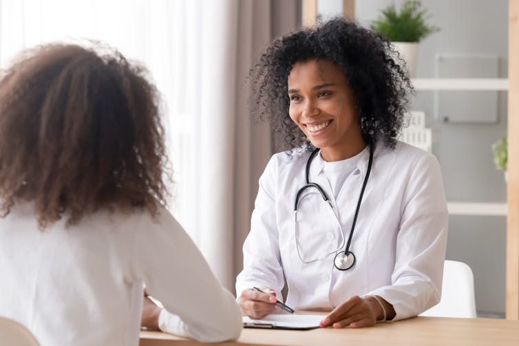 A woman in a white coat and stethoscope smiling as she talks to a young woman who is seen from behind