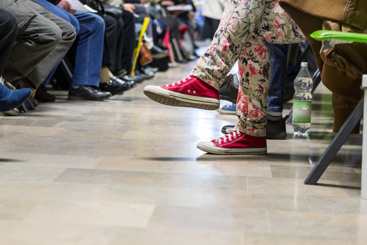 Feet of many people seated in a waiting room, with someone in red sneakers and flowered jeans in the foreground and many with brown and black shoes in the background.