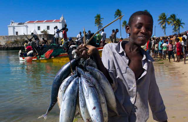 A young man carries fish as fishermen unload a boat in the background on a beach in Mozambique.