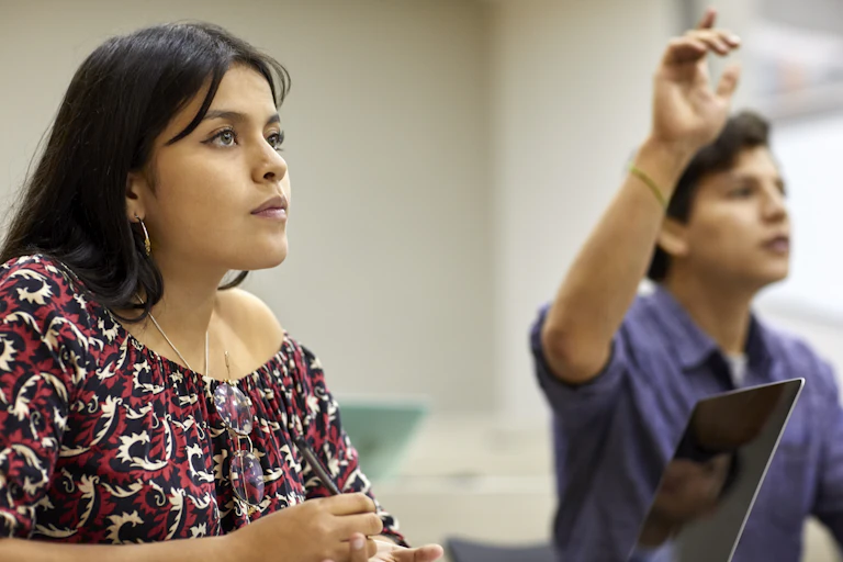 A female and male student sit at desk in a class and look at their instructor, who is unseen.
